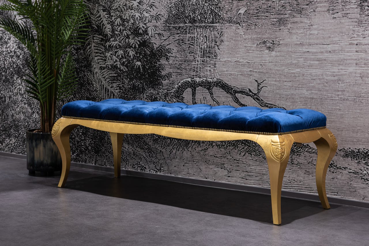 services-01 A stylish blue velvet bench with gold legs in a sophisticated interior setting with decorative wallpaper.